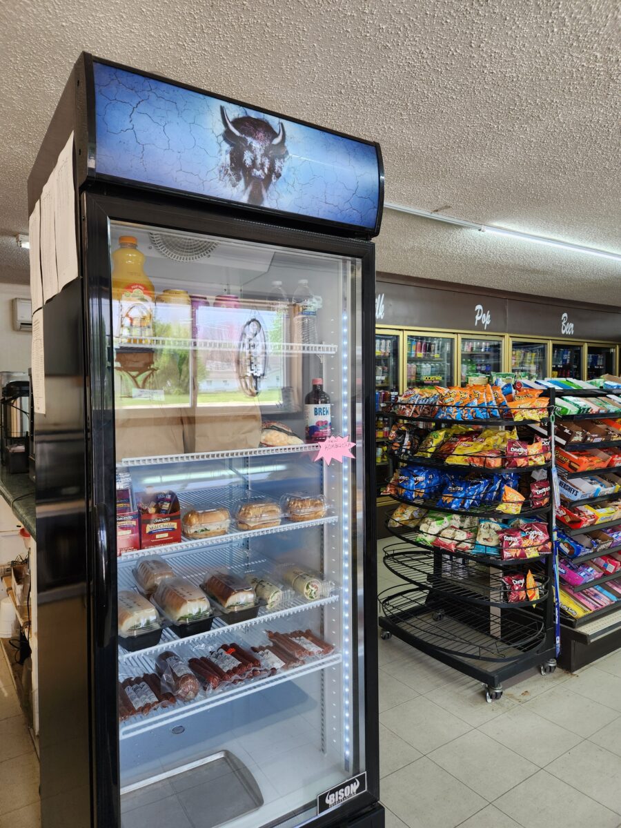A refrigerated display case in a store features various snacks. The top shelf has bottles of orange juice and a small Santa Claus figure. The middle and lower shelves have muffins, pastries, and packaged beef jerky. Adjacent shelves hold assorted chips and snacks.