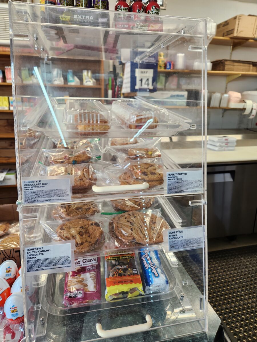 A clear display case on a countertop showcasing assorted cookies, muffins, and pastries. The case has several shelves with individually packaged treats, including peanut butter cookies, chocolate chip cookies, and bear claws. A calendar displaying the date 