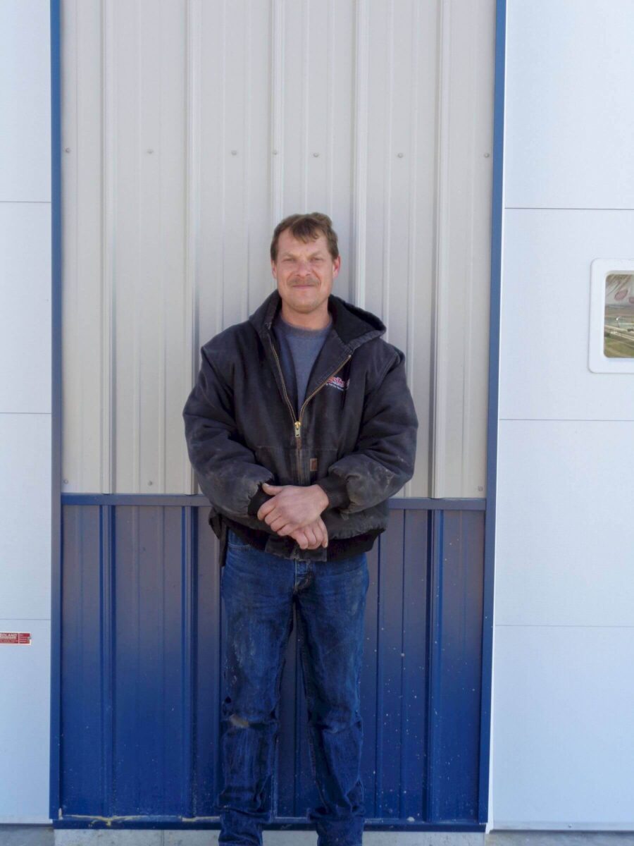 A man stands smiling in front of a corrugated metal door. He is wearing a dark jacket, blue jeans, and has his hands clasped in front of him. The background features blue and white panels.