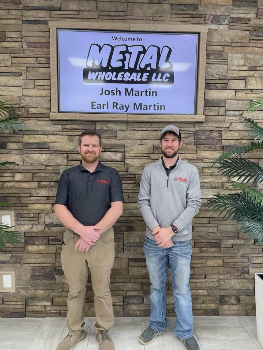 Two men are standing in front of a brick wall under a sign that reads 