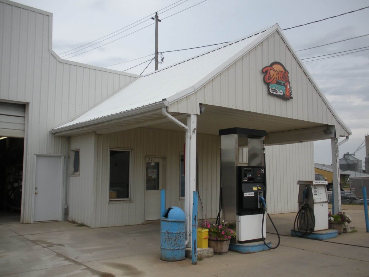 A small, white metal-clad gas station with a single fuel pump and a blue trash can out front. The building has a covered entryway with a sign above the pump reading 