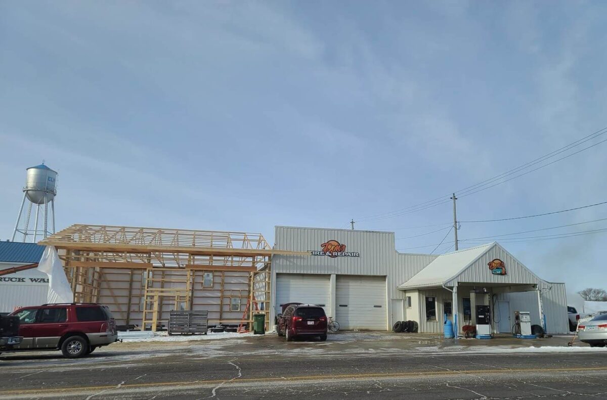A small service station with a white exterior sits under a clear sky. Vehicles are parked in front, and a partially constructed wooden structure is adjacent to the station. A water tower stands in the background. Signs on both buildings feature a colorful logo.