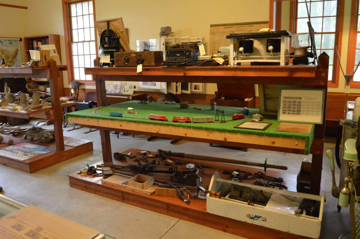 A room filled with historical artifacts displayed on wooden tables and shelves. Items include typewriters, a model train set, rifles, and various small objects. The room has large windows and tan walls, giving it a bright and organized appearance.