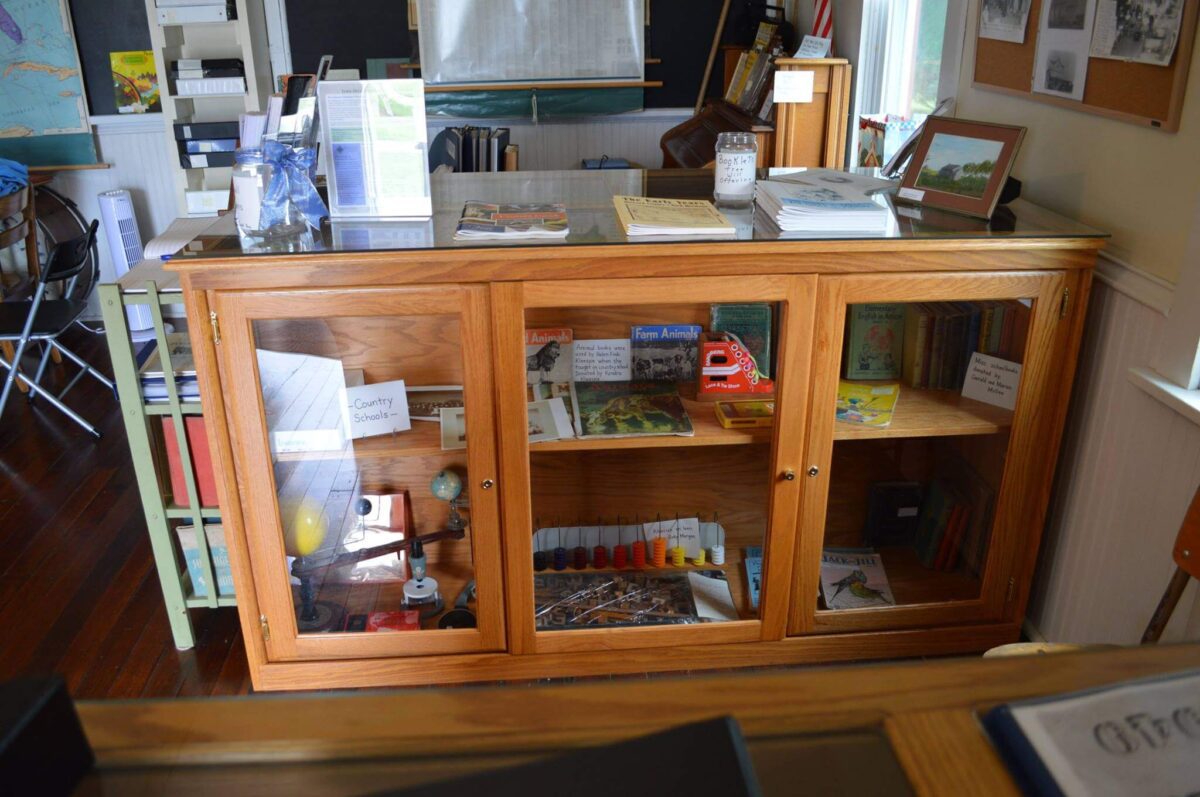A wooden display cabinet with three glass doors showcasing various books, bird figurines, and craft supplies like yarn and a color palette. The cabinet is placed against a wall in a well-lit room with additional books, brochures, and items on its top surface.