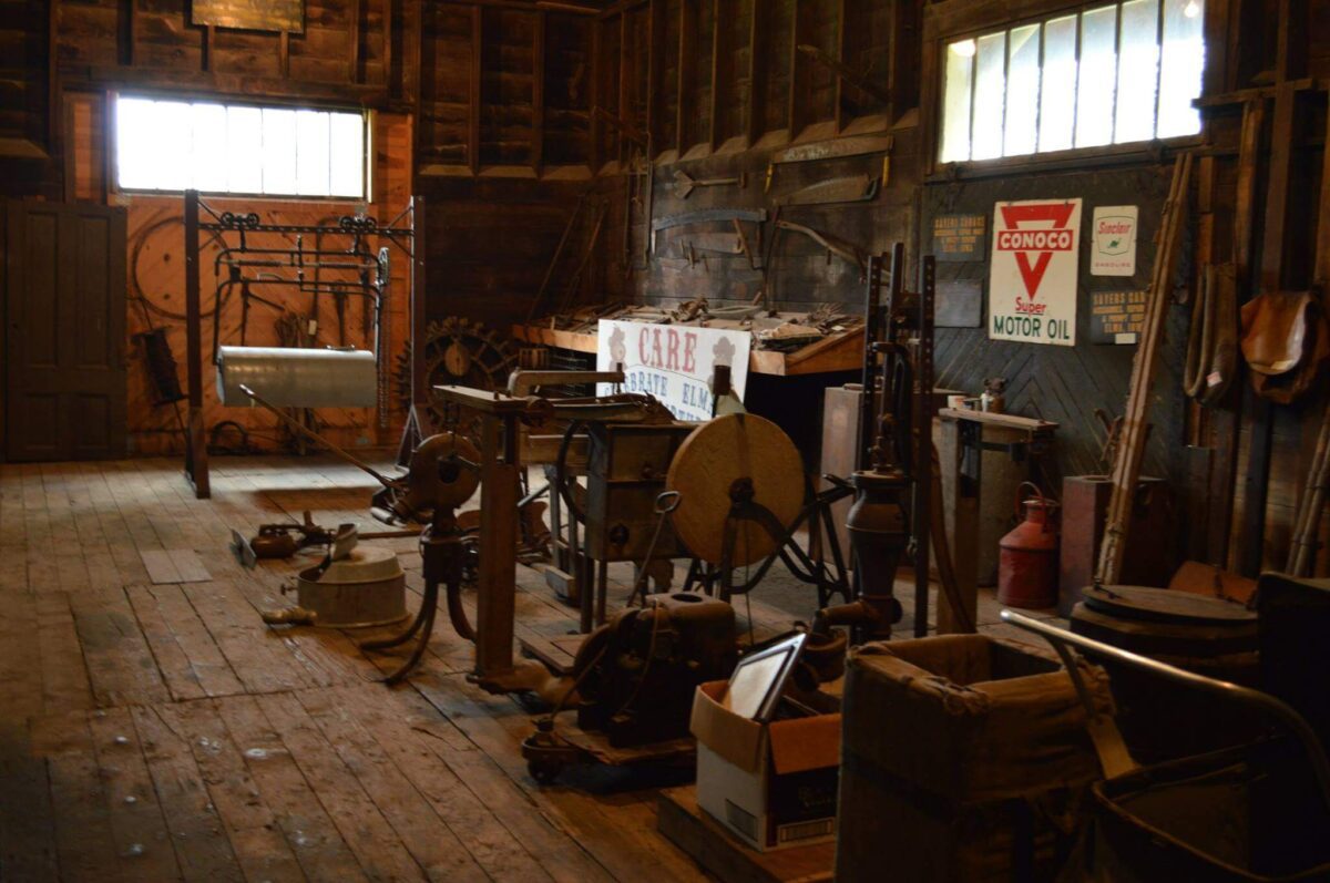 A dimly lit, rustic workshop with wooden floors and walls, containing various vintage tools and machinery. Signs, including one for Conoco Motor Oil, are mounted on the walls. A long workbench runs along one side, cluttered with tools and equipment.