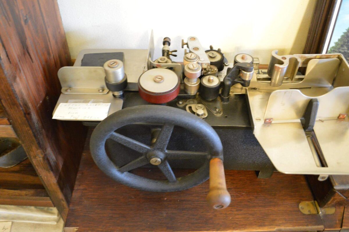 A vintage mechanical device featuring a large handwheel, several rollers, spools, and various metal components. It is mounted on a wooden surface, with a small slip of paper inserted into one side. The machine appears to be old and possibly used for printing or typing.