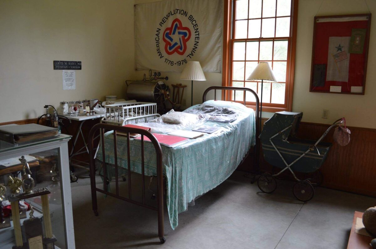 A room featuring an old-fashioned hospital bed with medical supplies, a vintage baby stroller, and historical artifacts. An American Revolutionary Bicentennial banner is on the wall. Sunlight streams in through the window, casting a warm glow over the scene.