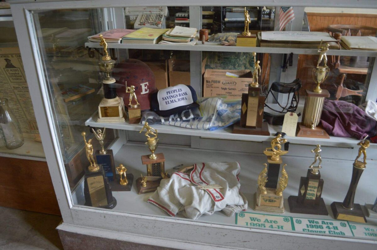 A display case filled with various trophies and awards is shown. The trophies vary in shape and size, with some resembling people in athletic poses and others with abstract forms. A few old newspapers and a red helmet with the letter 