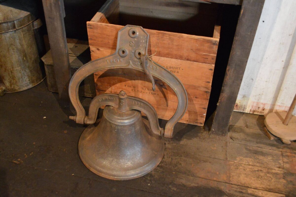 An old metal bell with a handle sits on a wooden floor in front of a wooden box. The bell has inscriptions and is part of a rustic, possibly historical, setting. The background includes various wooden and metal objects, adding to the vintage atmosphere.