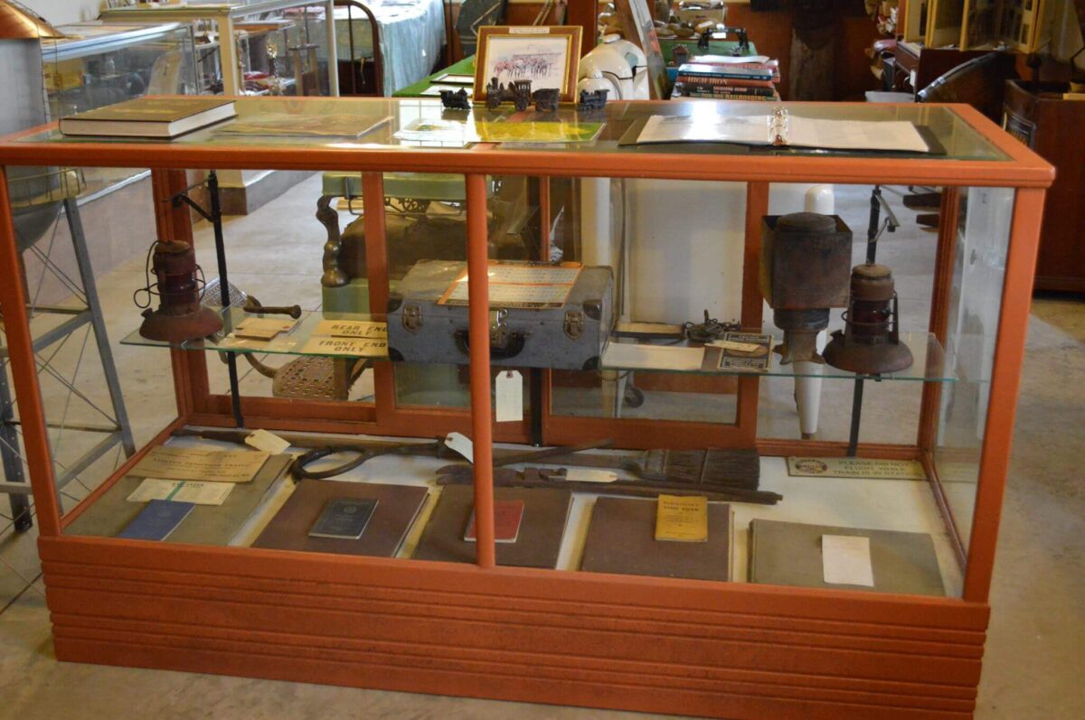 A glass display case with various historical artifacts, including vintage books, lanterns, a metal box, and tools. The case is situated in a spacious room with additional displays and items visible in the background.