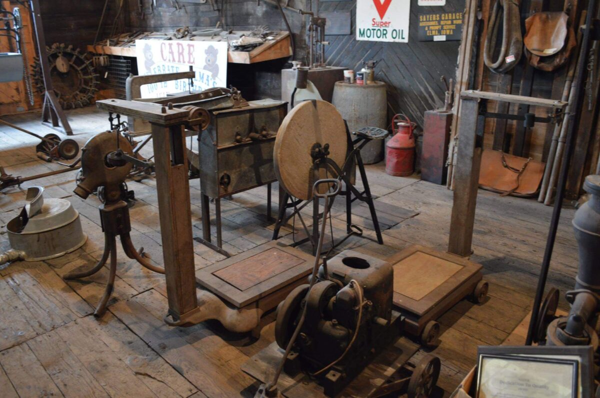 An old workshop filled with vintage machinery and tools. Various pieces of rusted equipment, wooden platforms, and a large grinding wheel are scattered around the wooden floor. A weathered sign for motor oil is visible on a corrugated metal wall in the background.