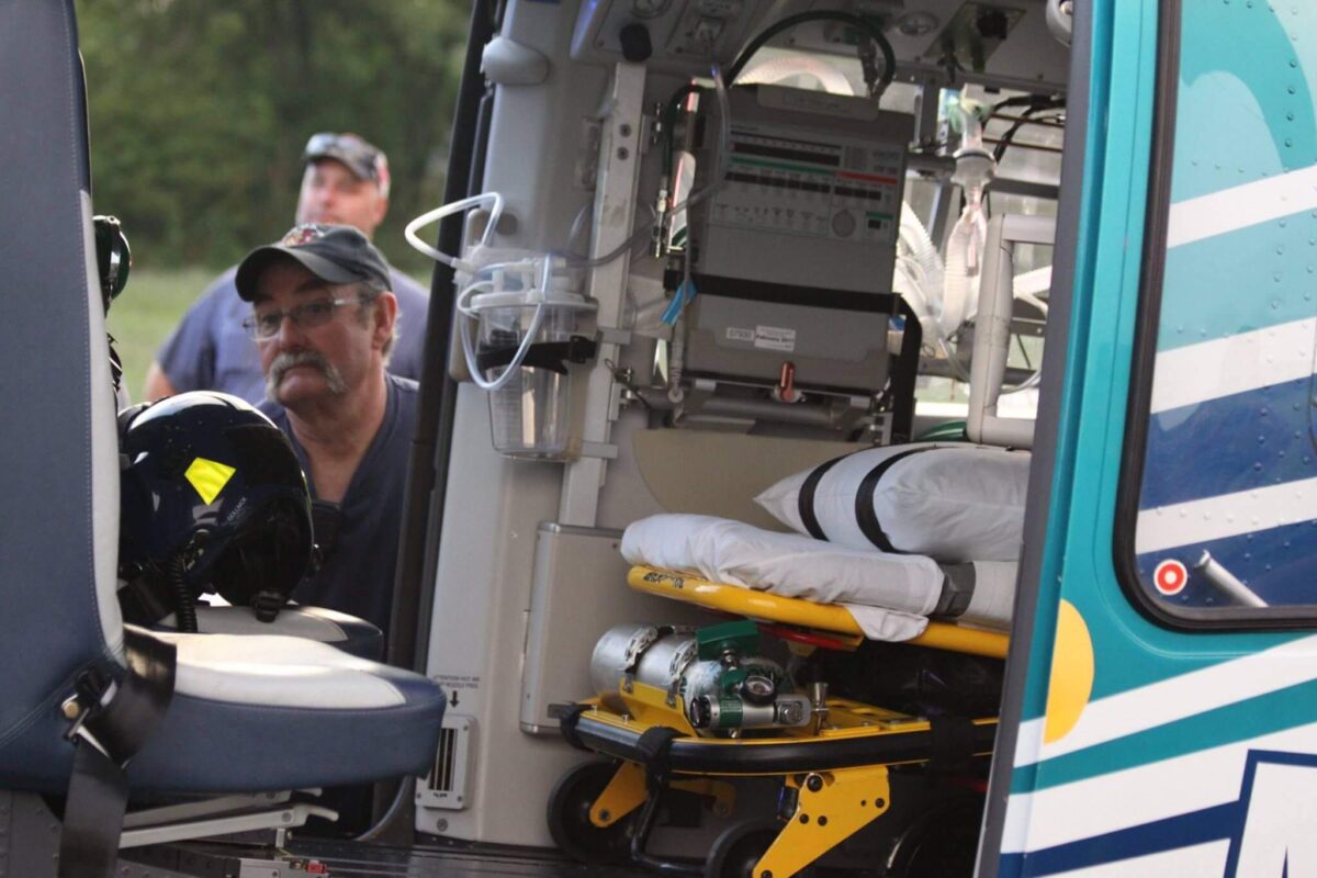 A man wearing a baseball cap and glasses looks into a helicopter's medical interior, which contains a stretcher with medical equipment and supplies. The helicopter's door is open, showing the detailed medical setup inside. Another person is visible in the background.