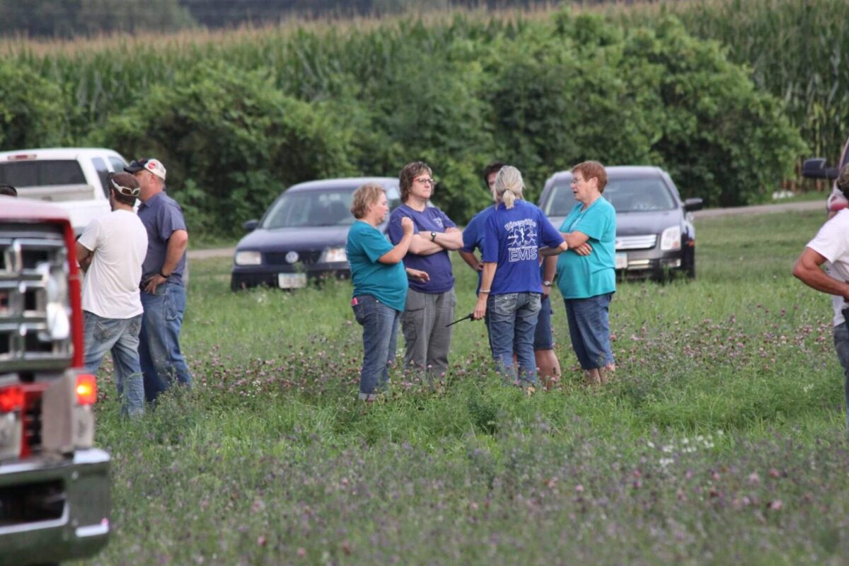 A group of people stand in a grassy field, some wearing colorful shirts. Several cars are parked in the background. The group appears to be having a conversation, and a fire truck is partially visible on the left side of the image. Trees and crops are in the background.