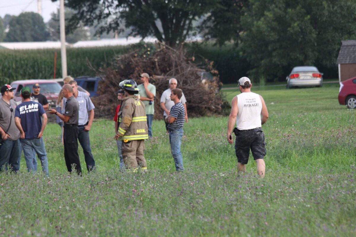 A group of people, including a firefighter in gear, stand in a field having a conversation. They are surrounded by grass and parked vehicles, with trees and a cornfield in the background. Another person walks away from the group, wearing a white tank top and camo shorts.