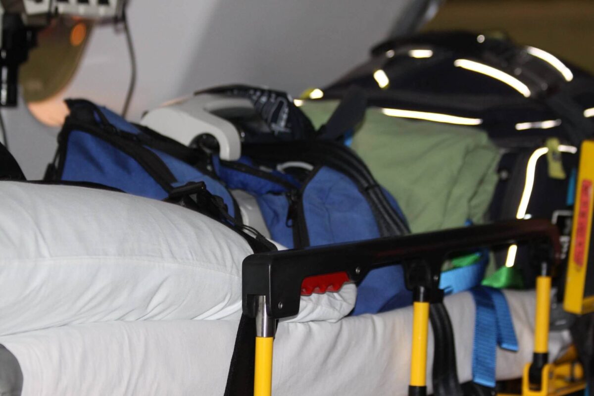 A close-up view of medical equipment and bags on an ambulance cot, including a blue bag, a green bag, and a white pillow. Yellow handles on the cot are visible in the foreground.