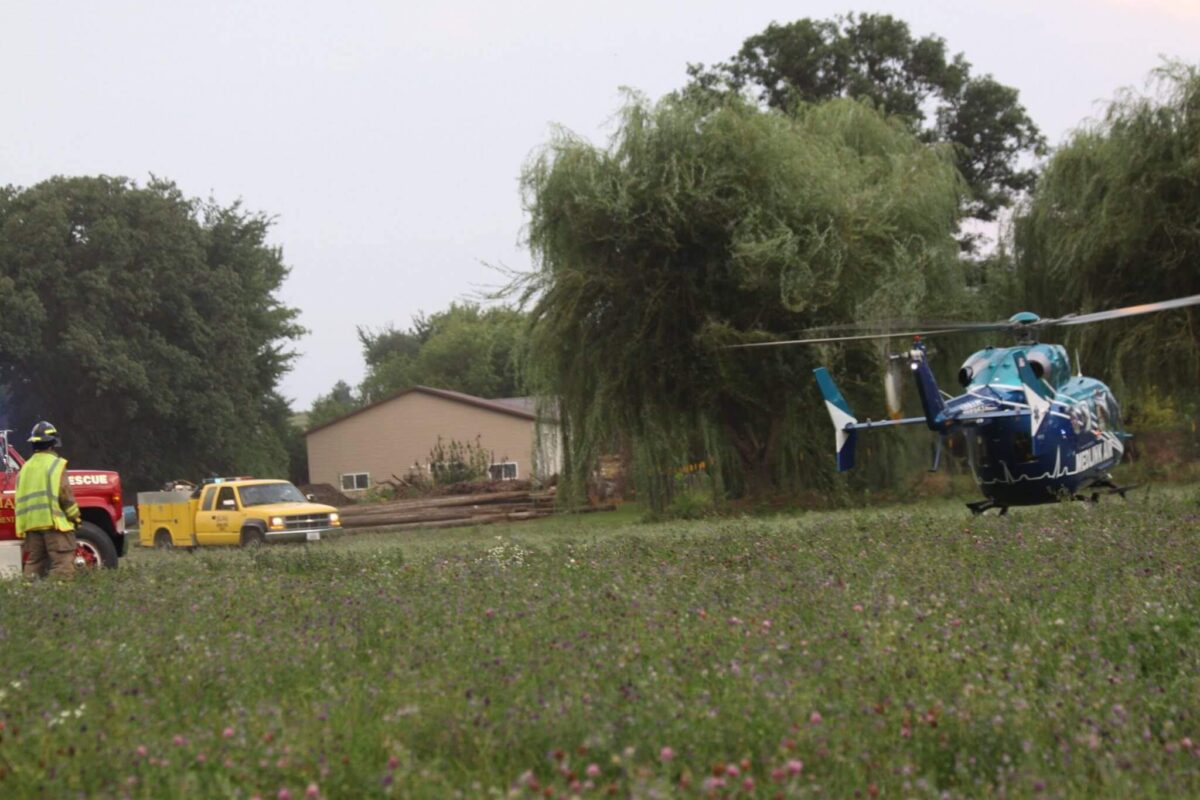A helicopter is landing in a grassy field with a rescue worker and emergency vehicles nearby. The worker is wearing a bright yellow jacket and a hard hat. Trees and a house are visible in the background.