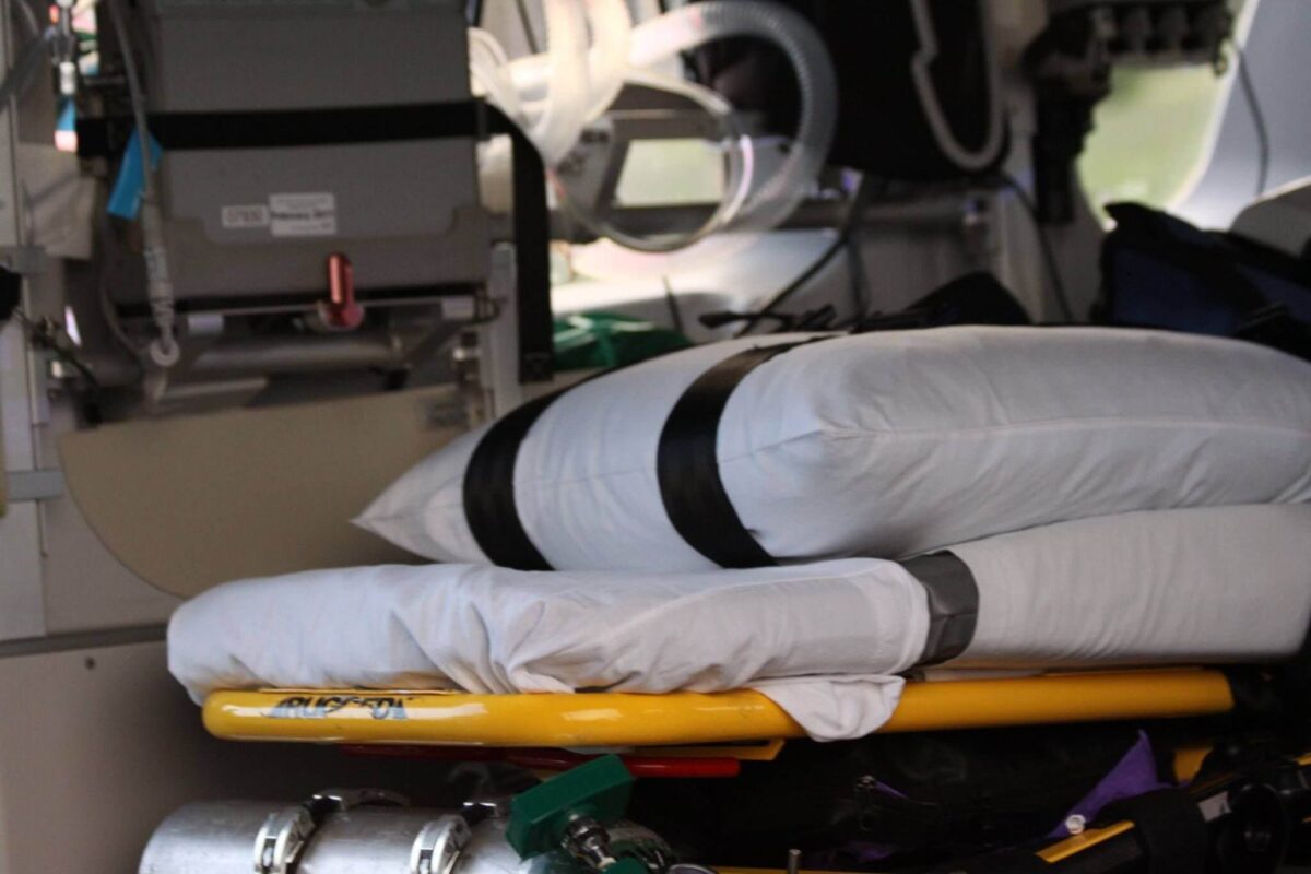 A medical stretcher on a helicopter is shown. The stretcher has pillows strapped on it. There are medical devices and equipment surrounding the stretcher inside the helicopter.