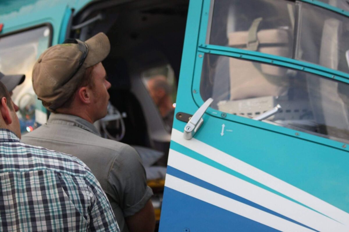 Two individuals stand near an open helicopter door. One person wears a beige cap and grey shirt, and the other wears a checkered shirt. The helicopter's exterior is blue with white and light blue stripes. The interior of the helicopter is partially visible.