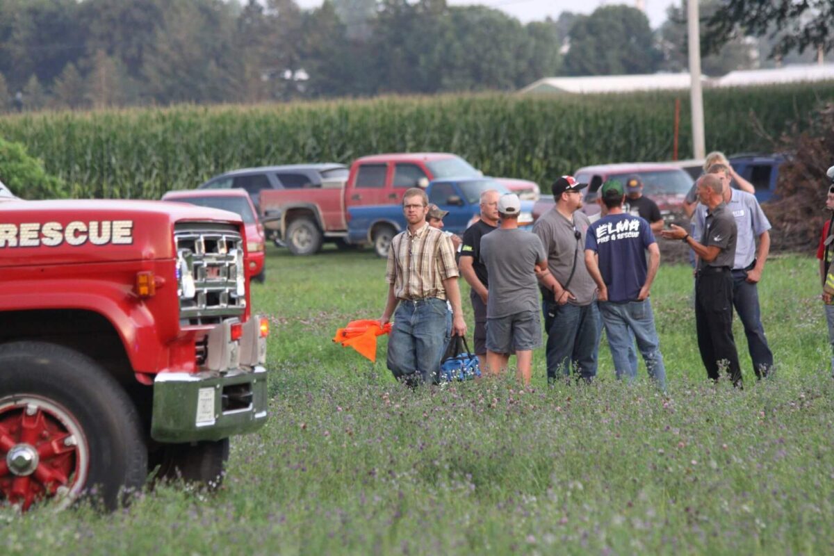 A group of people stands in a grassy field near a parked red rescue truck. Several individuals wear casual attire, while two wear emergency responder uniforms. In the background, there are multiple parked vehicles and trees.