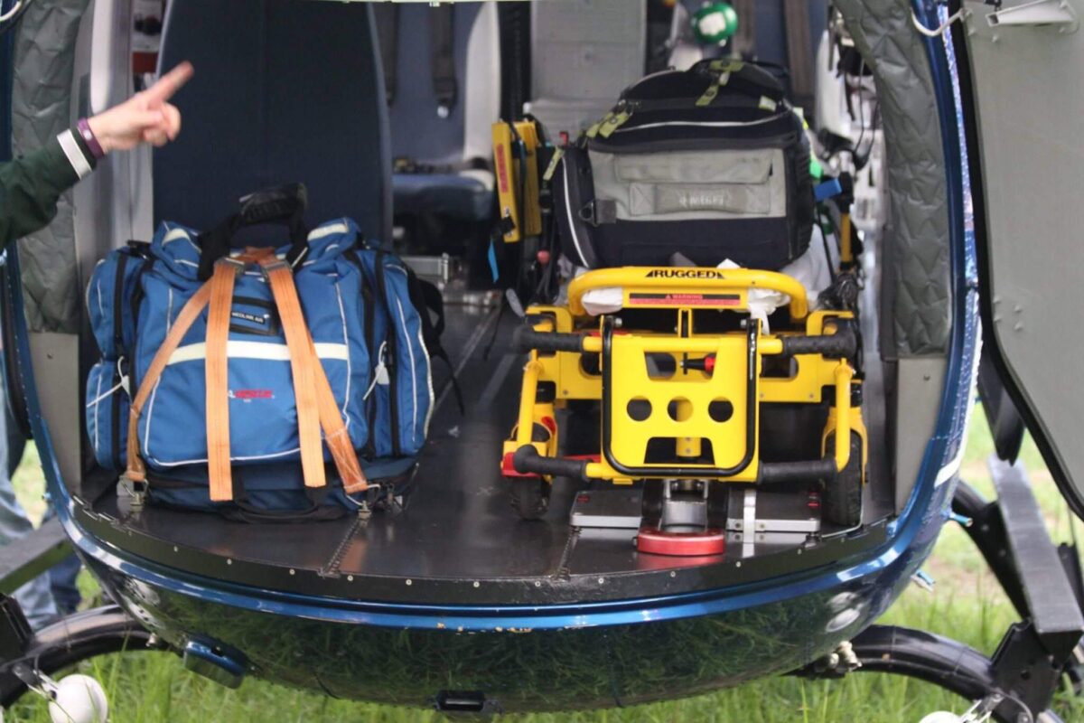Inside view of a rescue helicopter with a blue bag on the left and a yellow and black stretcher secured on the right. A person’s hand is visible on the left, pointing towards the bag. The helicopter doors are open, revealing grassy ground outside.