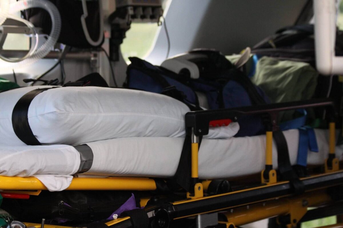 A close-up view of the interior of an emergency medical vehicle, showing a stretcher with a white pillow secured with black straps. Medical bags and equipment are visible in the background, suggesting preparedness for patient transport and care.
