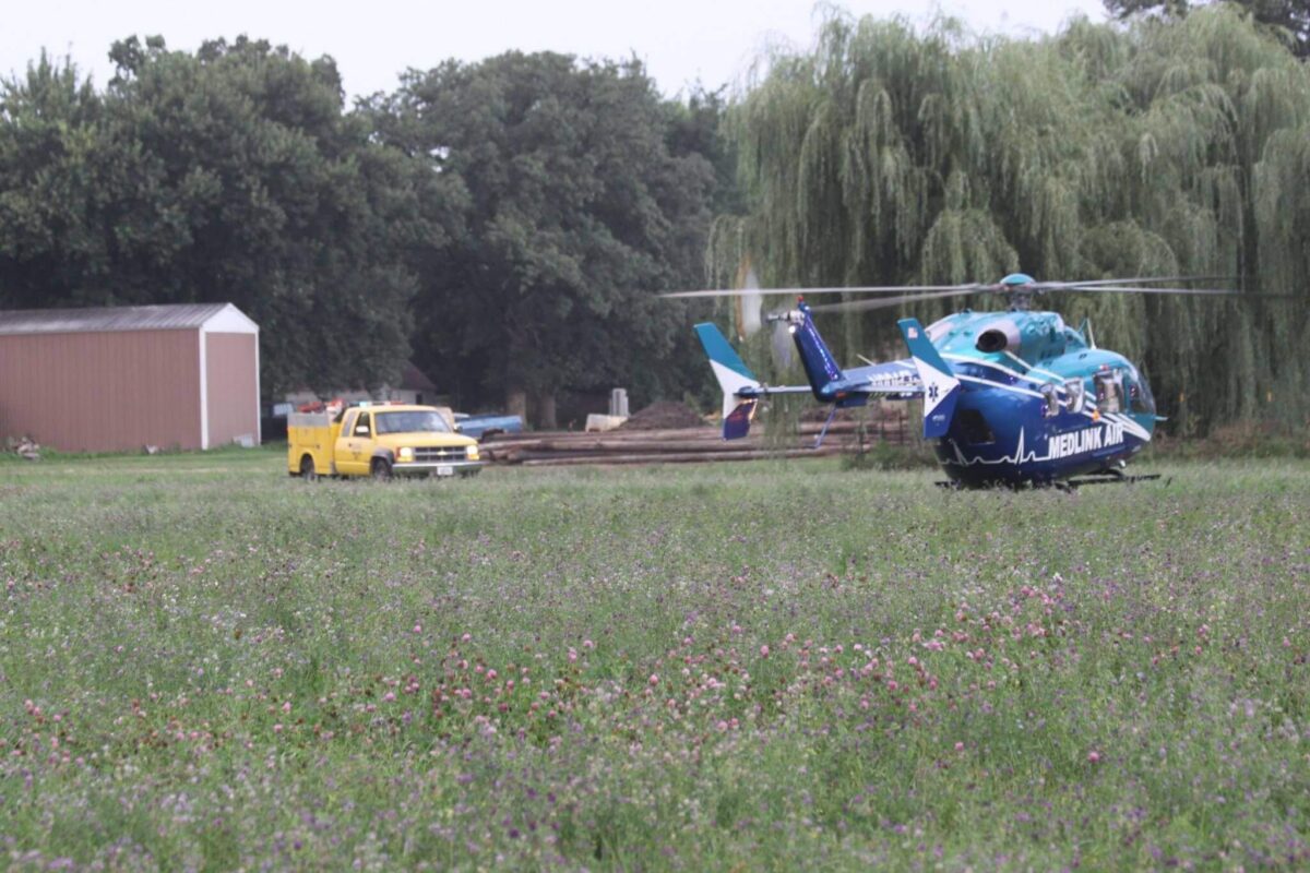 A blue and white medical helicopter is grounded in a field covered in low greenery and purple flowers, with a large willow tree and a yellow utility truck in the background. A barn stands nearby, partially obscured by trees.