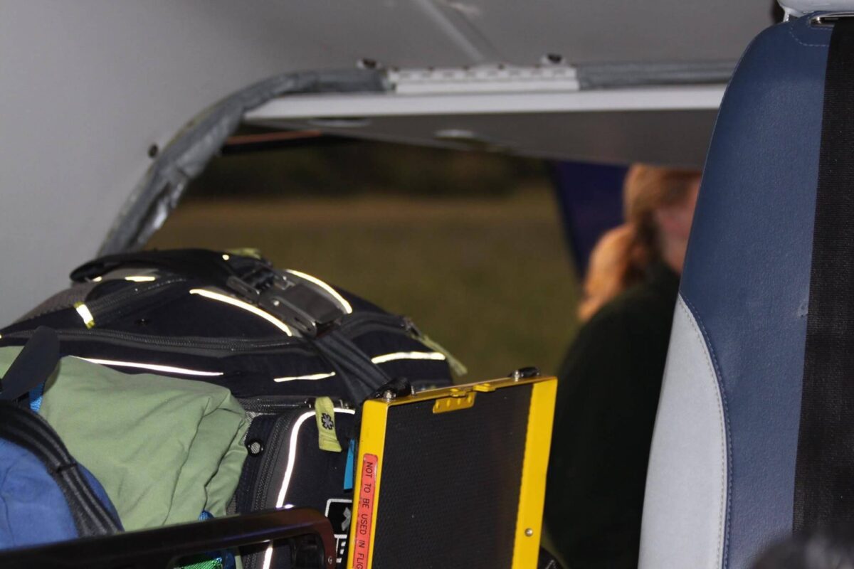 A close-up shot inside a vehicle, possibly an ambulance or rescue vehicle. The focus is on emergency gear and equipment stacked on a seat, including reflective strips and a clipboard. A blurred figure is visible in the background through an open door.