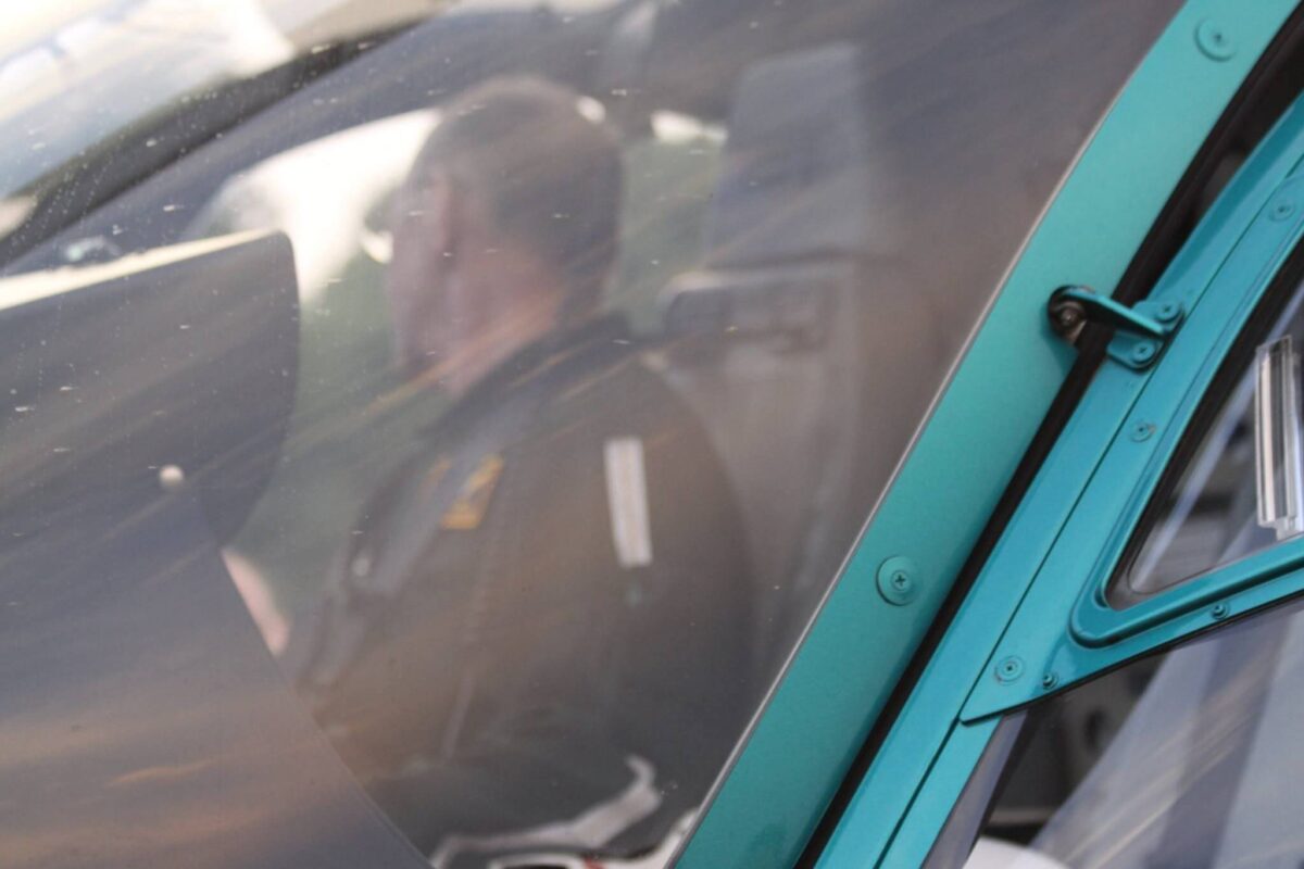Close-up view through a helicopter window showing the silhouette of a person seated inside. The image is somewhat blurry, emphasizing the perspective from outside the helicopter, with the window framing part of the interior.