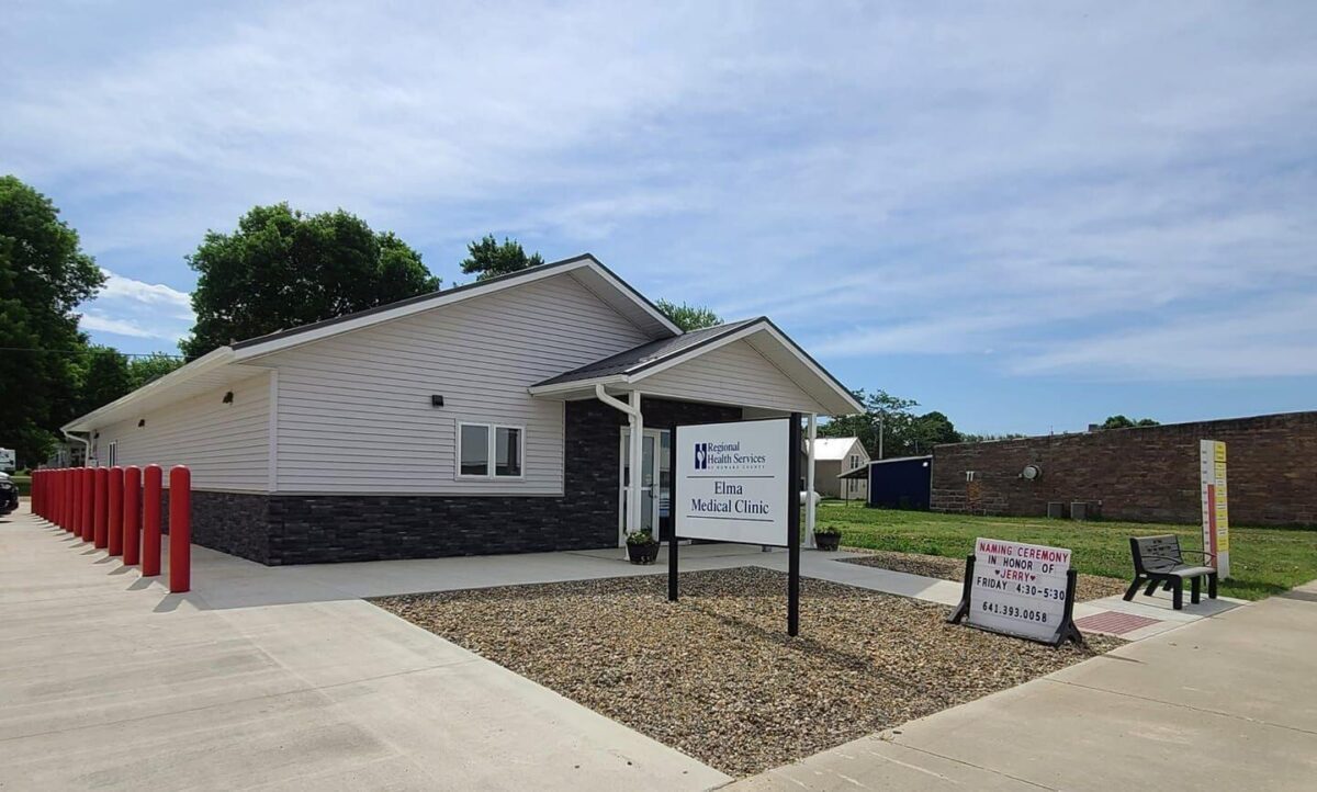 A single-story white building with a dark gray brick base houses the Etna Medical Clinic. A sign in front bears the clinic’s name and hours. Red bollards line the sidewalk to the left, and a small bench is positioned near another sign on the right. Trees and a parking lot are in the background.