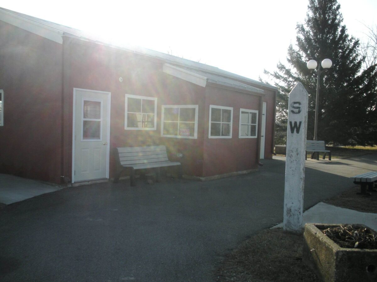 A small red building with several windows and a white door is seen in sunlight. A bench is placed to the left side of the door. A tall concrete post marked 