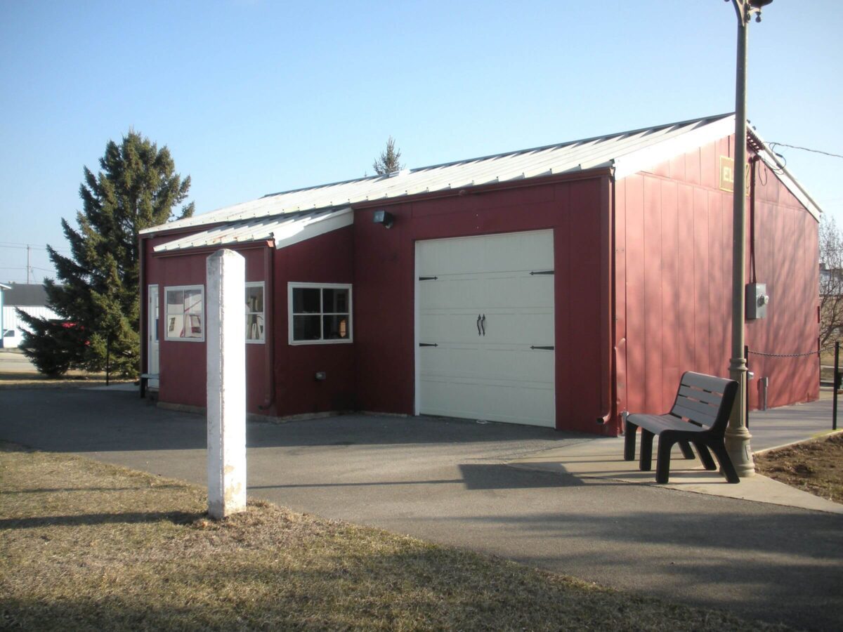 A red building with a white garage door and two windows on its side. There is a black bench on the right, near a street lamp. The area is paved, with grass on either side and a large tree in the background.