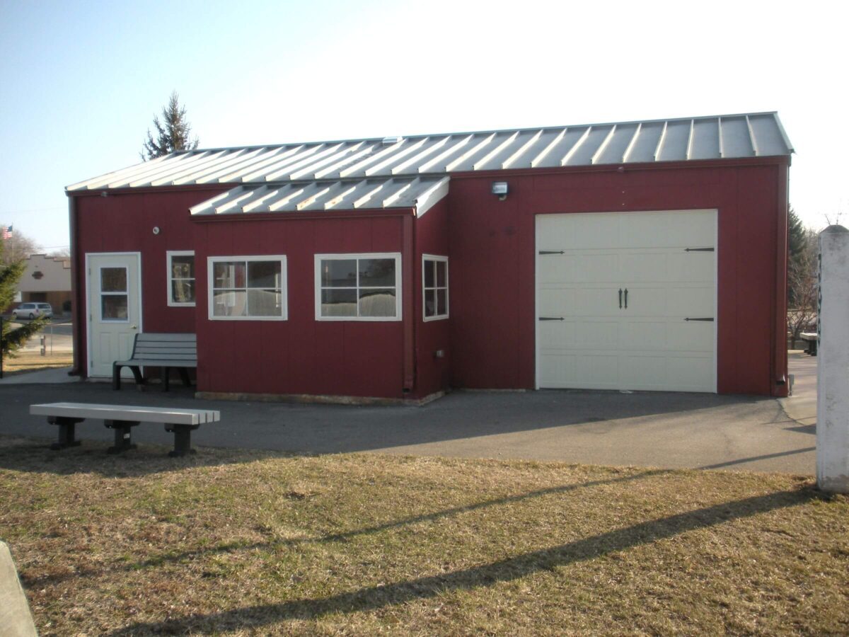 A small, red-painted building with a metal roof and multiple windows. It has a white garage door and two regular doors. Two benches are placed outside on a paved area next to the building. The surrounding area has grass and a few trees.