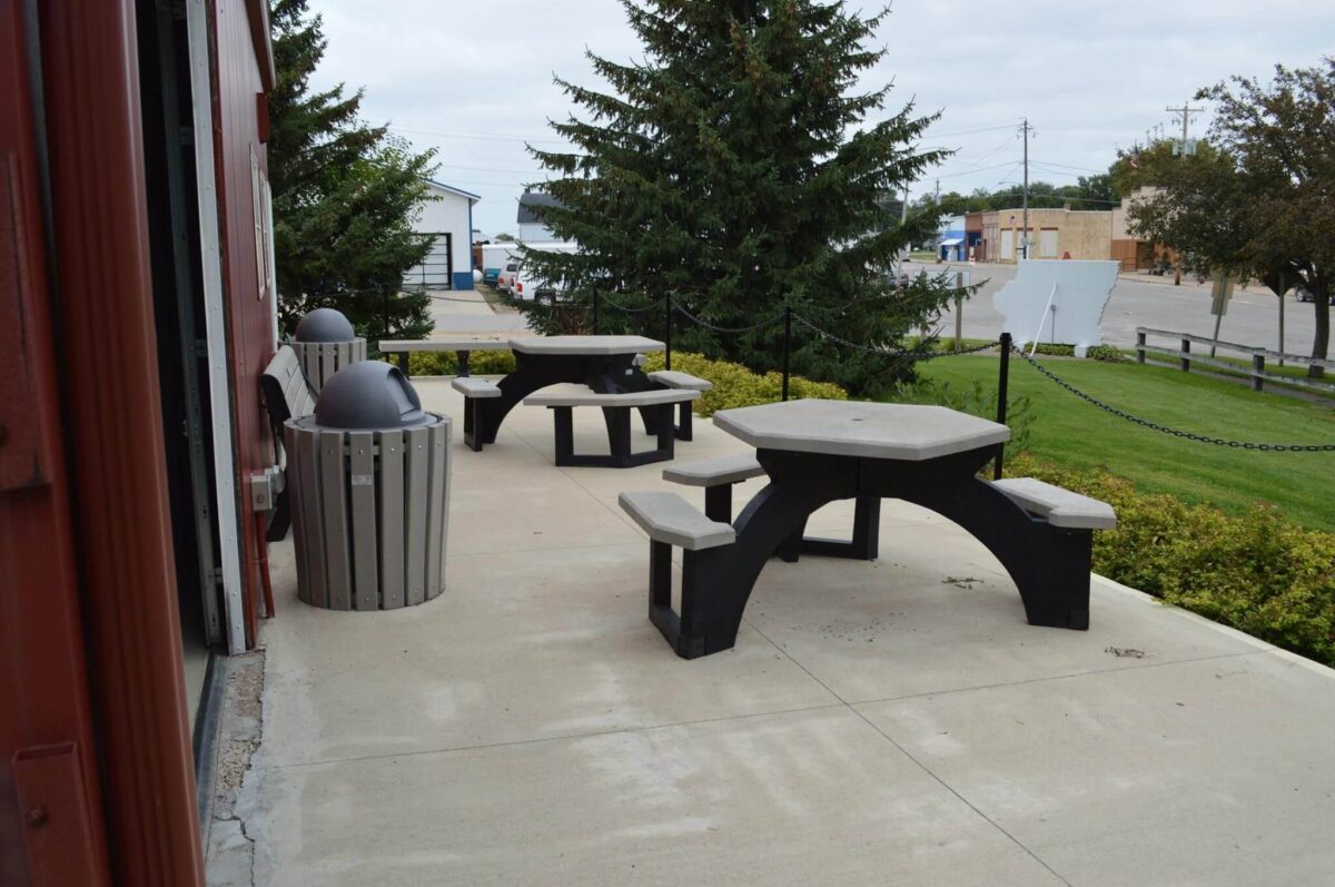 Outdoor patio with two black tables and benches beside a building. Two round trash bins with dome lids are nearby. Trees, bushes, a chain link fence, and a distant view of buildings and a parking lot are visible in the background. Sky is overcast.