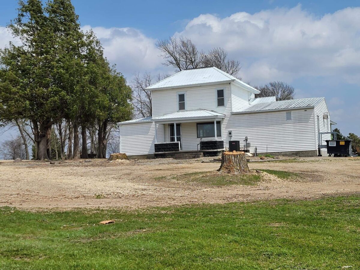 A white, two-story house with a metal roof is situated on a large, mostly barren plot of land with tree stumps scattered around. A group of tall trees stands to the left of the house. The sky is partly cloudy. A black dumpster is near the right side of the house.