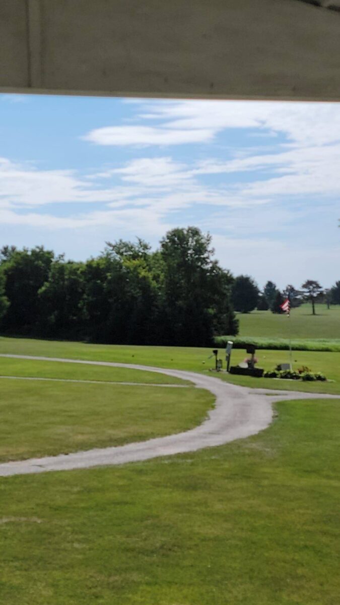 A winding paved path cuts through a well-manicured green lawn, leading towards a cluster of trees in the distance on a sunny day. An American flag stands near a small flowerbed along the path, and the sky is partly cloudy.