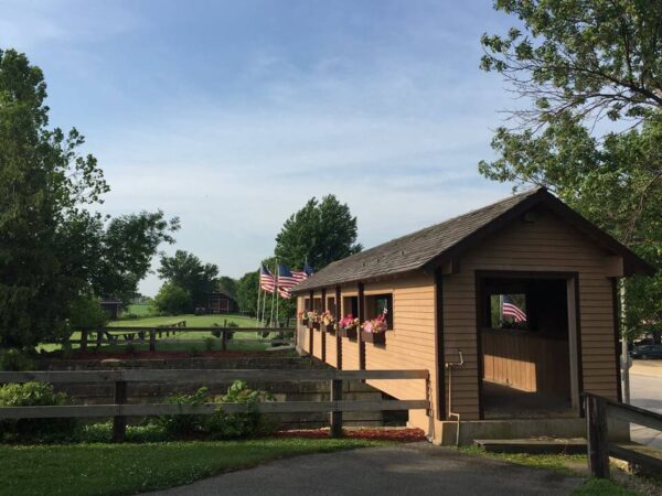 A wooden covered bridge adorned with hanging flower baskets and American flags. Trees and green grass surround the area, with a clear blue sky overhead. A pathway leads to the bridge, and a wooden fence lines the side.