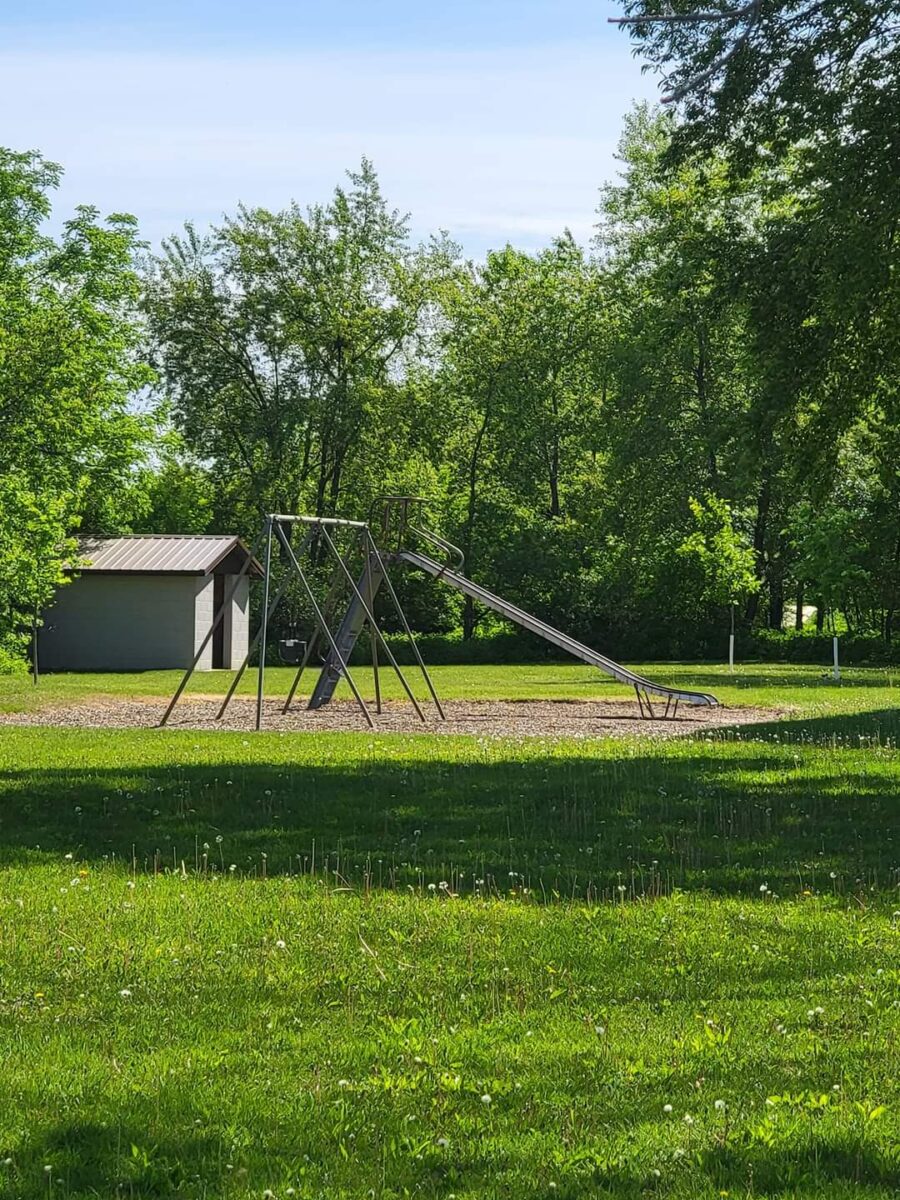 A playground with a metal slide and swings is surrounded by lush green grass and trees. A small shed is visible in the background, and the sky is clear and blue. The area is bright and sunny, creating a serene and inviting atmosphere.