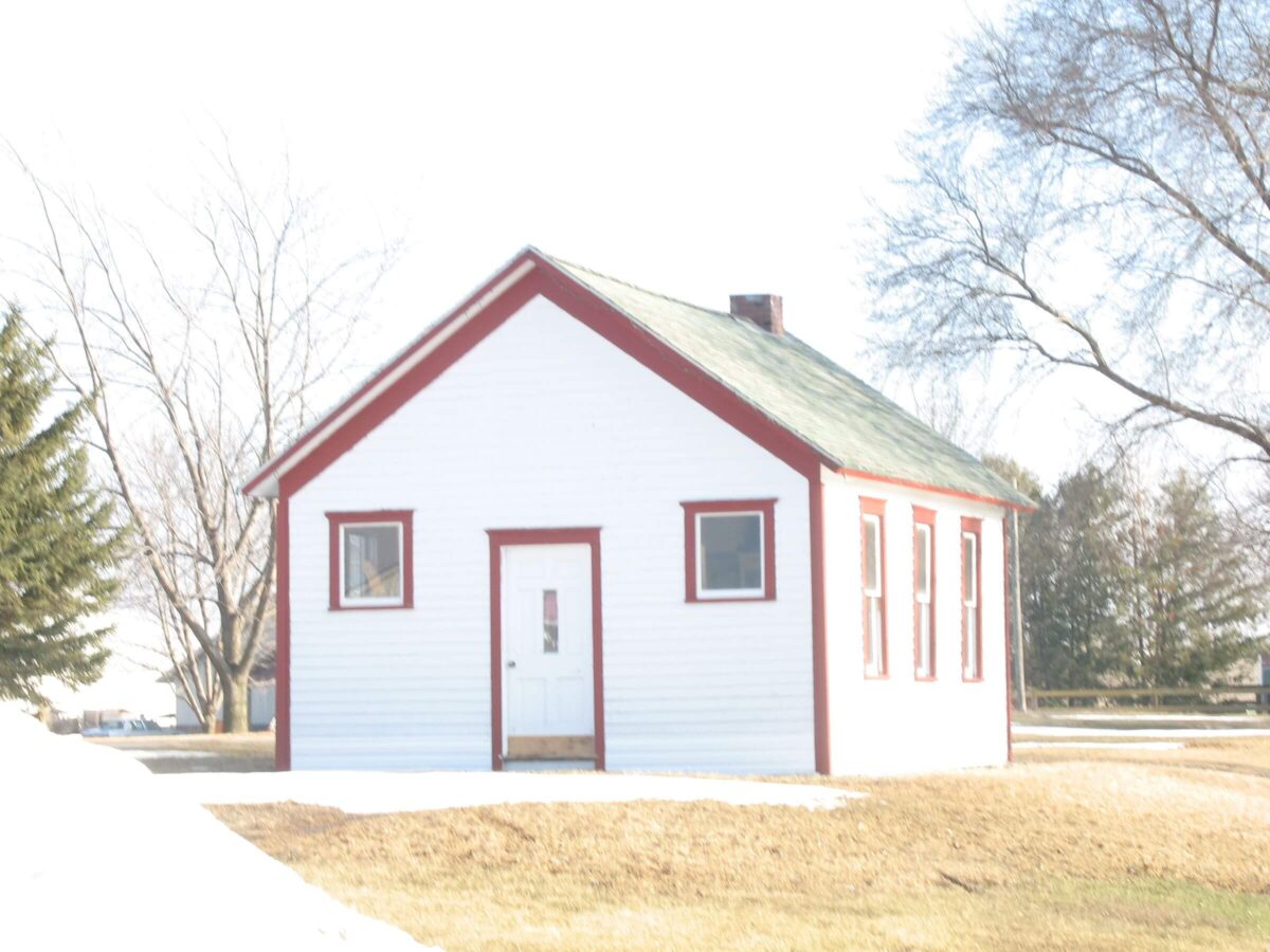 A small, white, rectangular building with a green roof and red trim stands surrounded by barren trees. The structure has a simple door on the front and multiple windows. The ground shows patches of dry grass and snow. The sky is clear and bright.