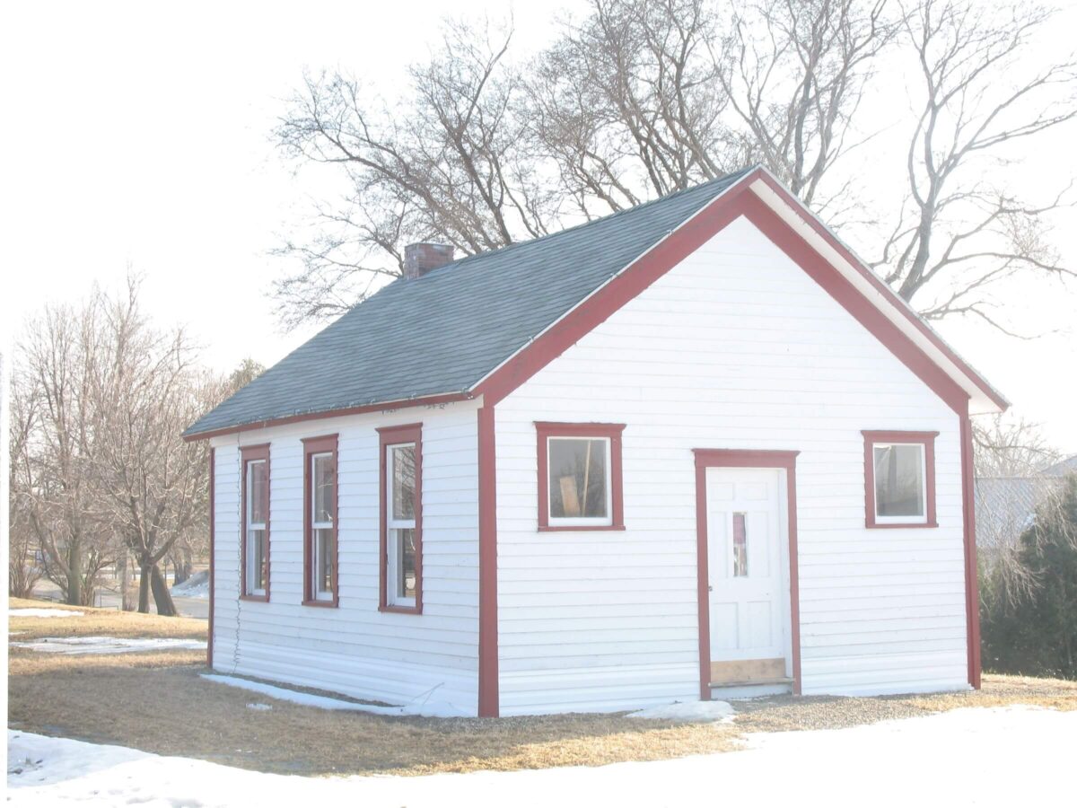 A small white house with a red-trimmed roof and windows is surrounded by bare trees and patchy snow on the ground. The house has three visible windows on one side and a central front door. The scene appears to be in a rural area on a sunny day.