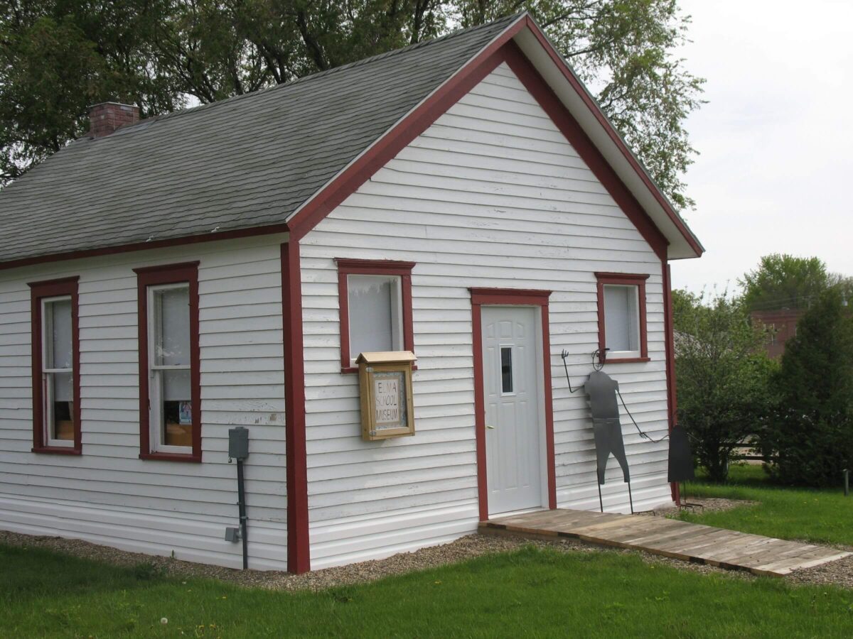 A small white wooden house with a gray shingled roof and red trim around the windows and door. A sign on the wall says 