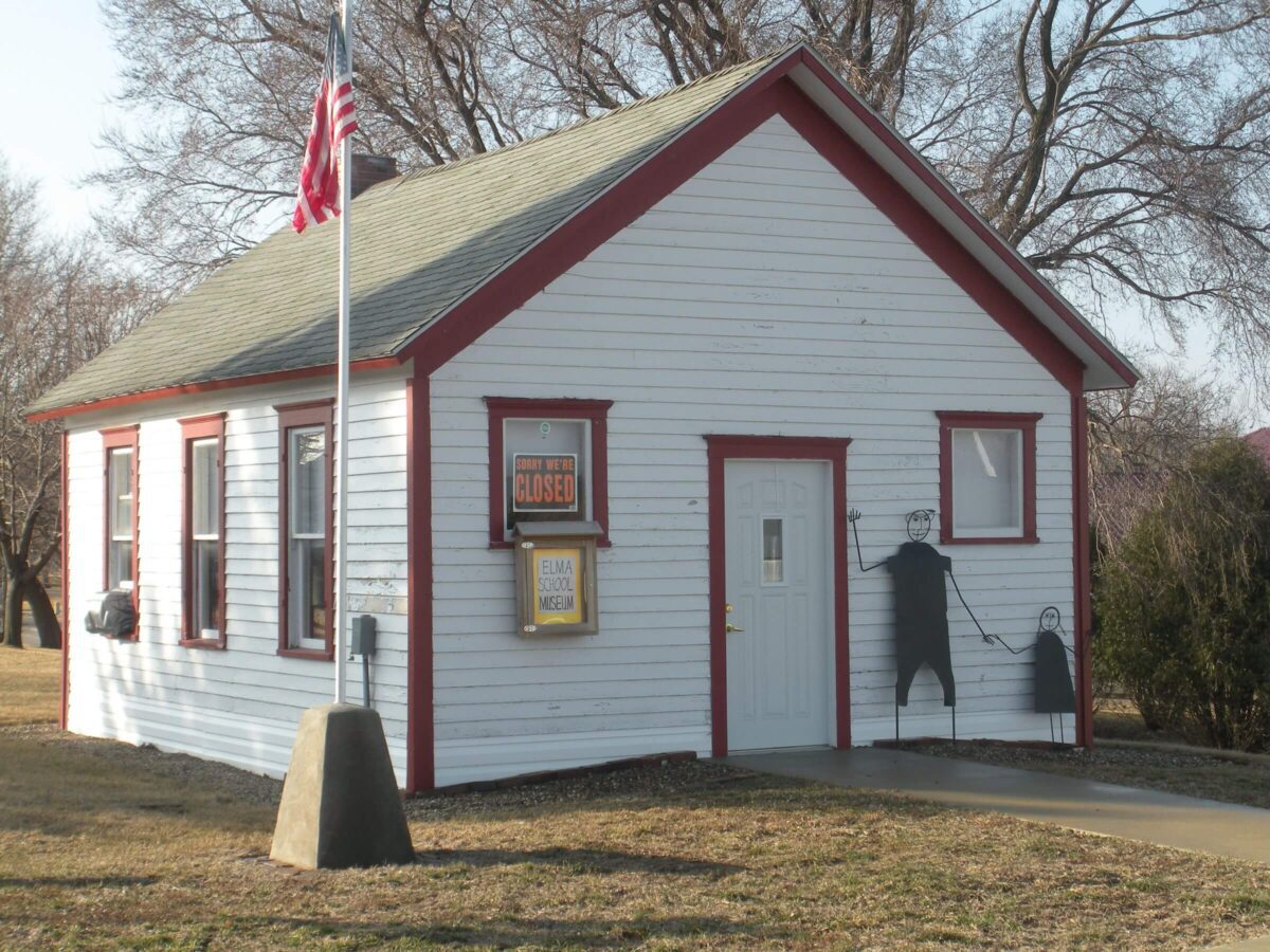 A small white wooden building with red trim, displaying a “CLOSED” sign and an American flag on a pole in front. Silhouettes of two children holding hands are painted on the side wall. Another sign reads 