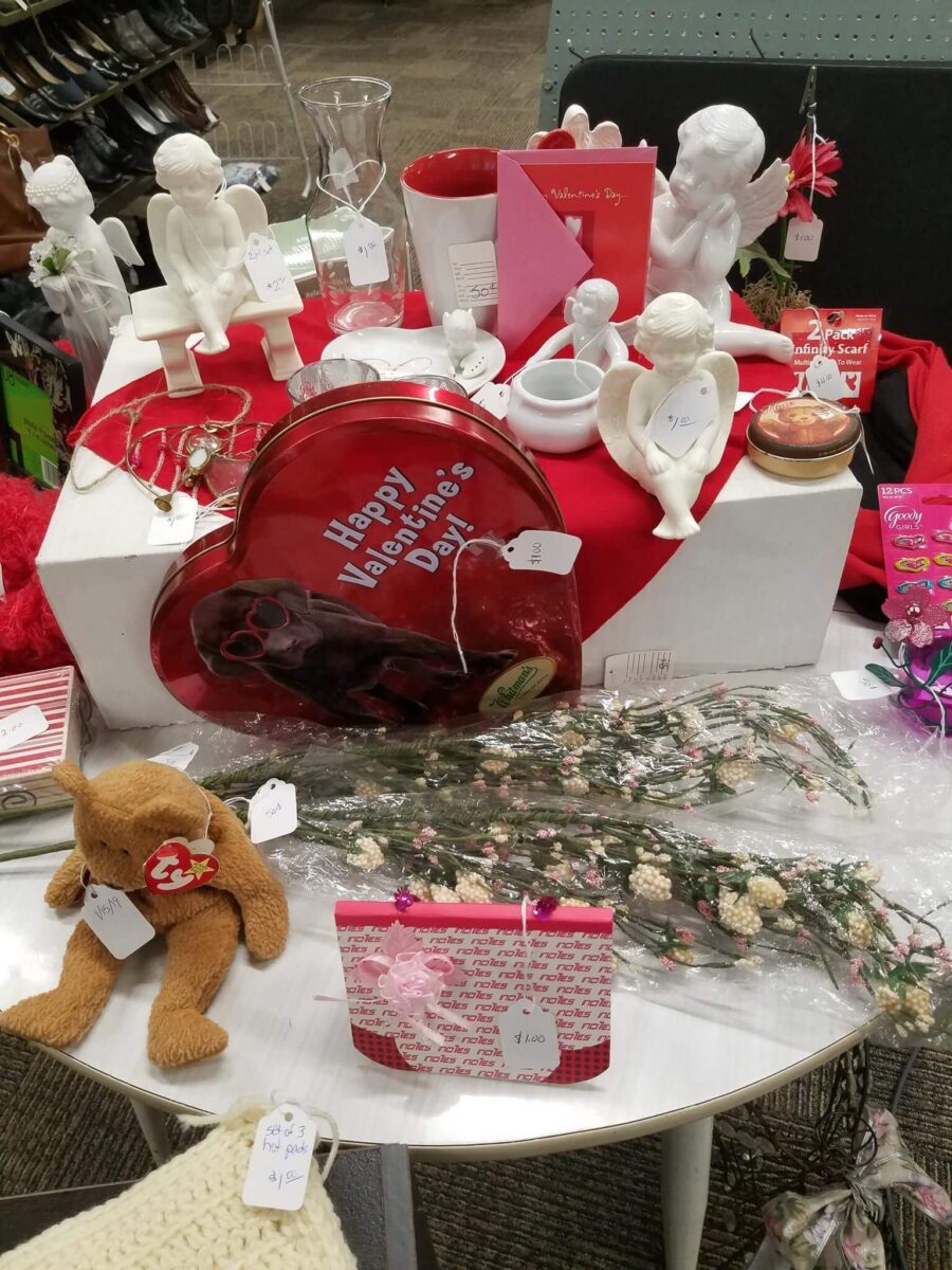 A display table showcasing various Valentine's Day-themed items, including heart-shaped boxes, ceramic angels, red and pink cards, a teddy bear, dried flowers, and other decorations. Each item has a price tag, and the setup is arranged on a white surface.