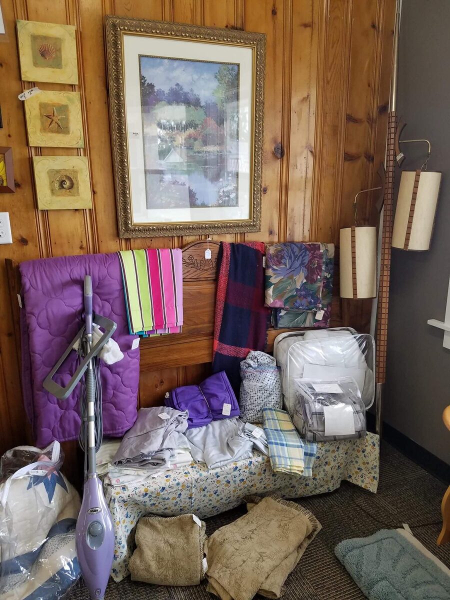 A cozy corner display featuring various home goods, including a purple vacuum cleaner, bed linens, towels, and patterned textiles. The wooden wall serves as the backdrop, adorned with framed art and decorative elements. A floor lamp with three lampshades stands to the right.