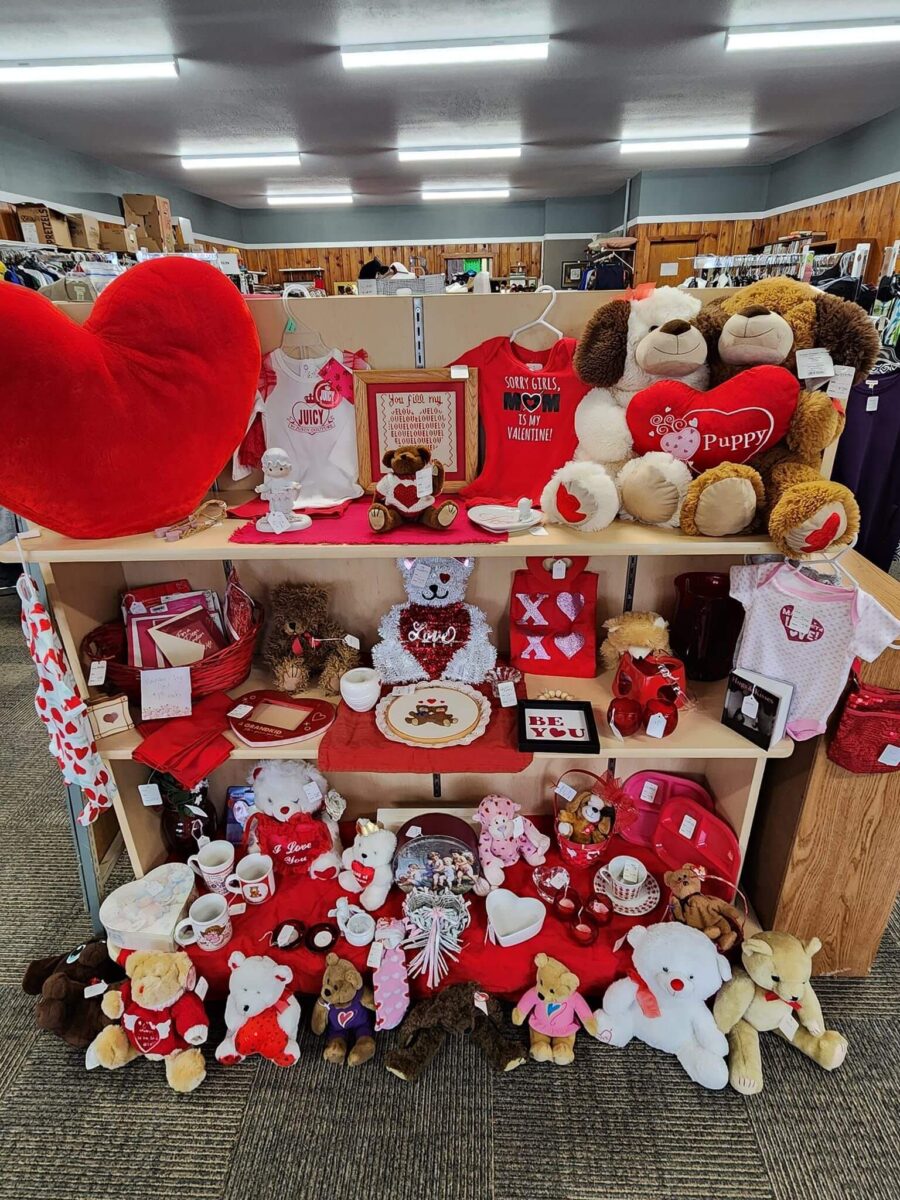 A store display featuring Valentine's Day-themed items, including plush toys, heart-shaped decorations, mugs, and clothing. The shelves are adorned with red and white accents, and various teddy bears and stuffed animals convey a romantic theme.