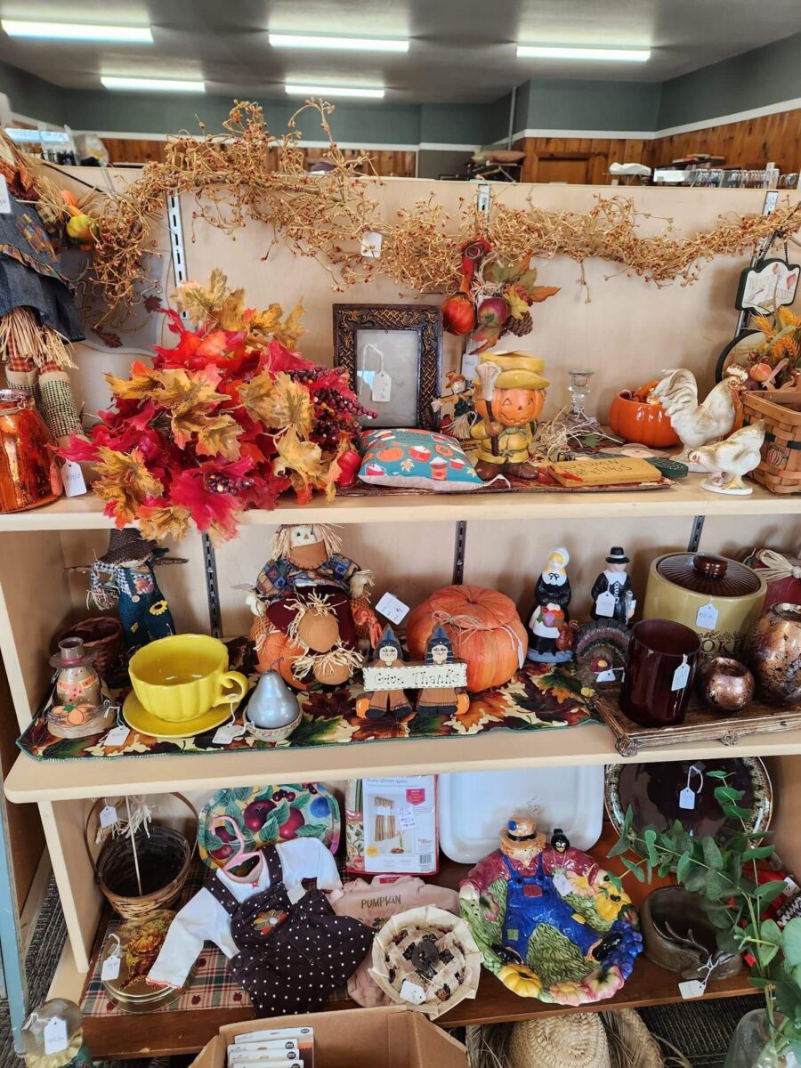 A store display of autumn-themed items, including scarecrows, pumpkins, fall leaves, and decorative figurines. The shelves feature a variety of ceramics, candles, and kitchenware adorned with seasonal motifs, arranged on a backdrop of orange and brown foliage.