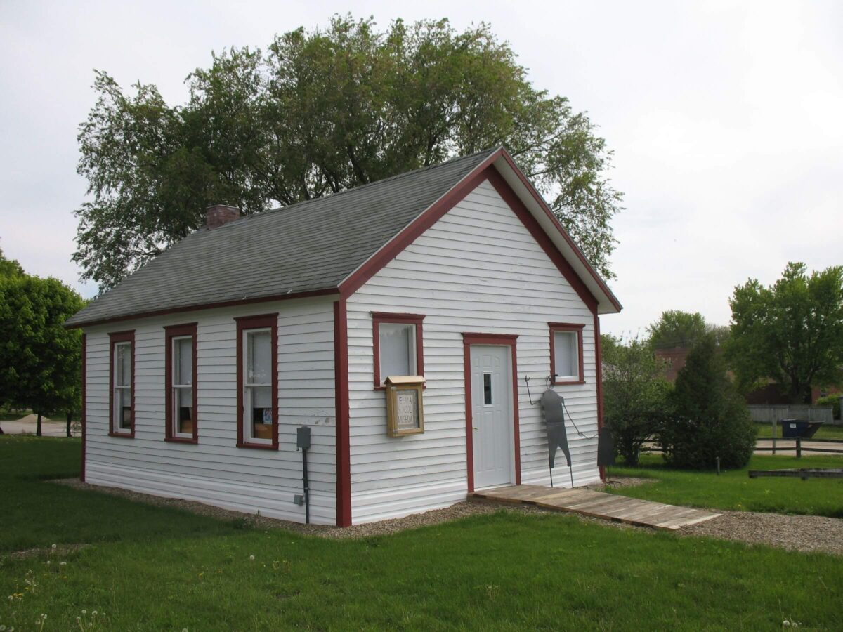 A small white wooden house with a gray roof and red trim sits on a grassy lawn. It has four windows, two on each side, and a single door in the front. A wooden ramp leads up to the door. Trees and a fence are visible in the background.