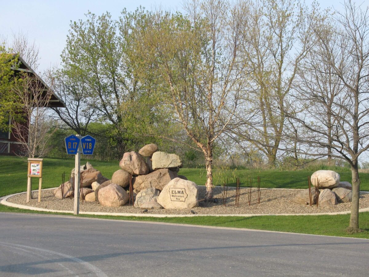 Landscape view featuring a roadside welcome sign for Elma. Surrounding the sign are large boulders and newly planted trees. A circular, paved drive surrounds the display. In the background, there’s a small building and clear, sunny skies with sparse trees.