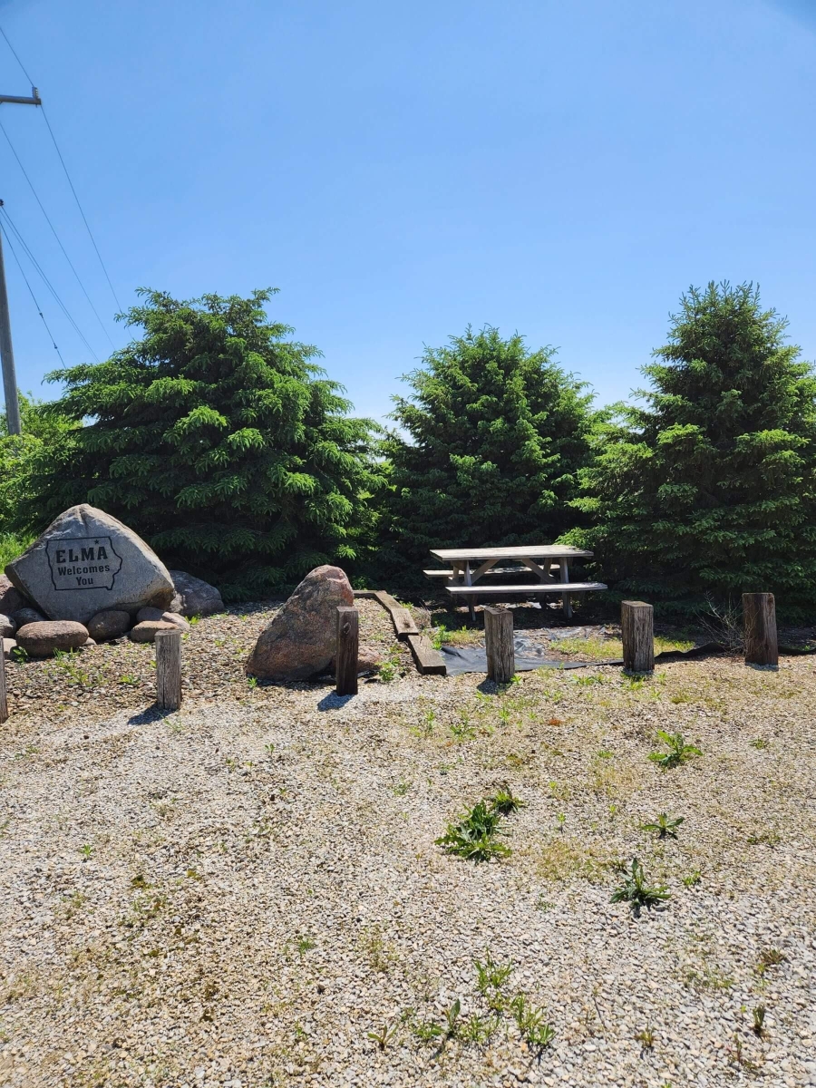 A small park area with a picnic table set between two large evergreen trees. A gravel path leads to the table, and a large rock inscribed with the word 