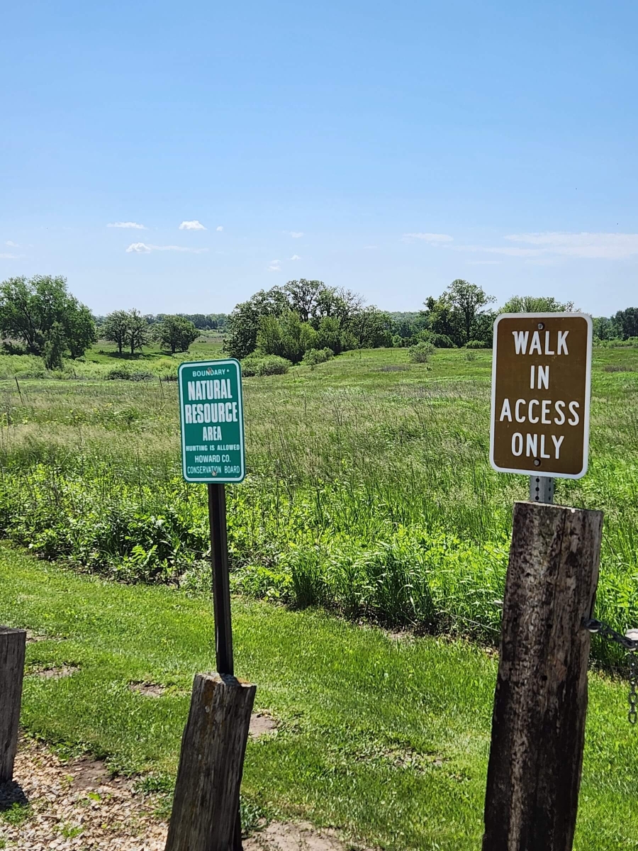 Two signs stand in front of a lush green field. The left sign reads 